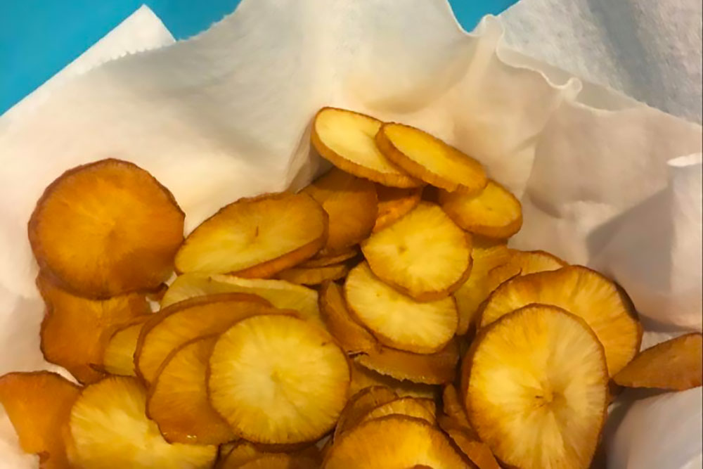 Homemade-Casava-Root-Chips A basket of golden-brown, crispy plantain chips is set against a blue background, showcasing a vibrant, allergen-friendly snack option. These natural chips are perfect for those seeking fresh flavors and healthy living.| Daré Vita Café | Las Vegas