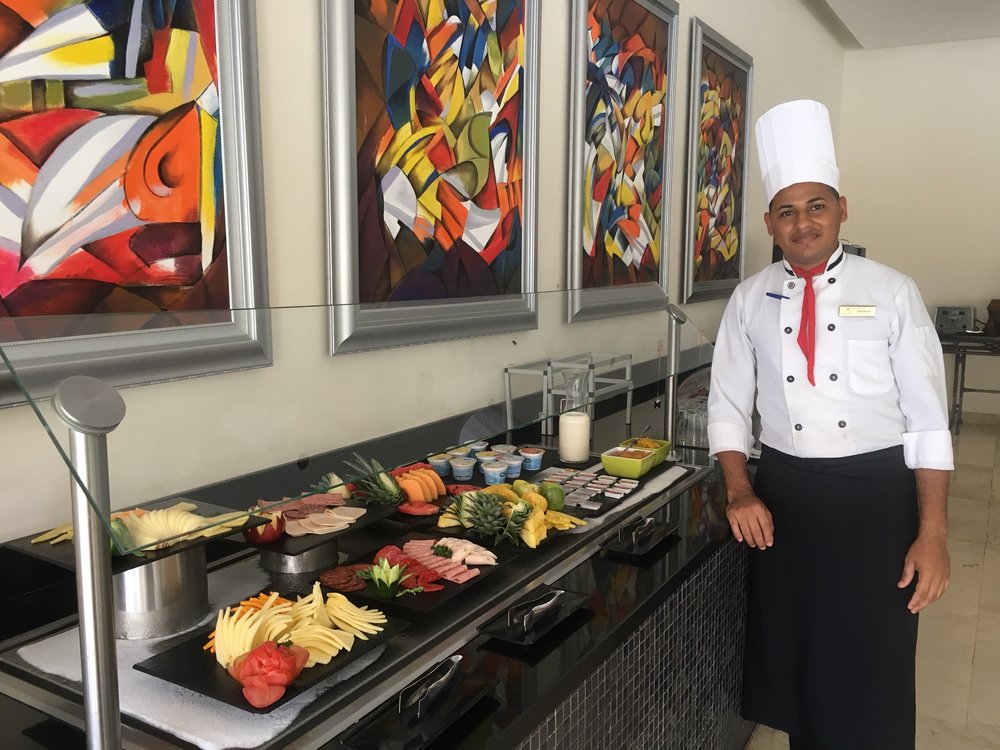 A chef in a white uniform stands beside a buffet table adorned with fresh produce and vibrant fruits, including pineapples and melons, under colorful abstract paintings, highlighting a focus on fresh, wholesome meals and healthy living.| Daré Vita Café | Las Vegas