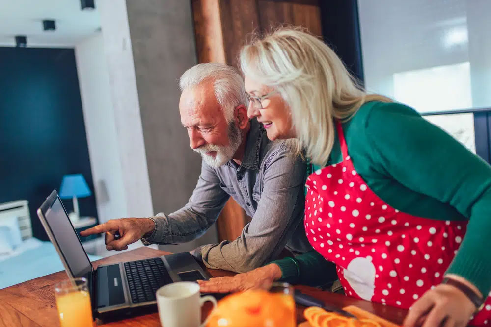 An elderly couple, with the woman in a red polka-dot apron, smiles while looking at a laptop in a cozy kitchen, featuring fresh orange slices, suggesting a wholesome and healthy living environment.| Daré Vita Café | Las Vegas