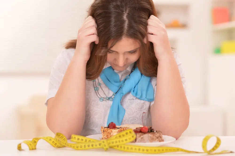 A woman with a blue scarf looks down at a cake on a plate, surrounded by a measuring tape, symbolizing food sensitivity. The scene highlights the importance of allergen-friendly and healthy living choices.| Daré Vita Café | Las Vegas