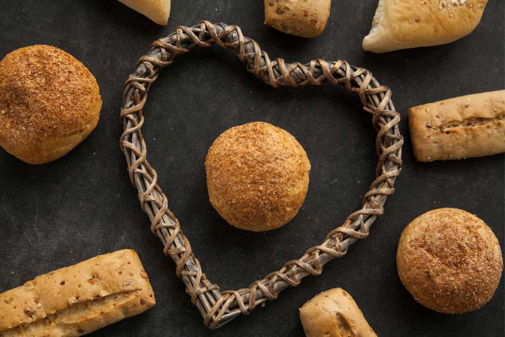 Embracing-a-Revolution-in-Healthy-Eating-1 A heart-shaped wicker frame surrounds a round, golden-brown bread roll on a dark surface, surrounded by various other breads. The image highlights the natural, wholesome ingredients used for allergen-friendly baking.| Daré Vita Café | Las Vegas
