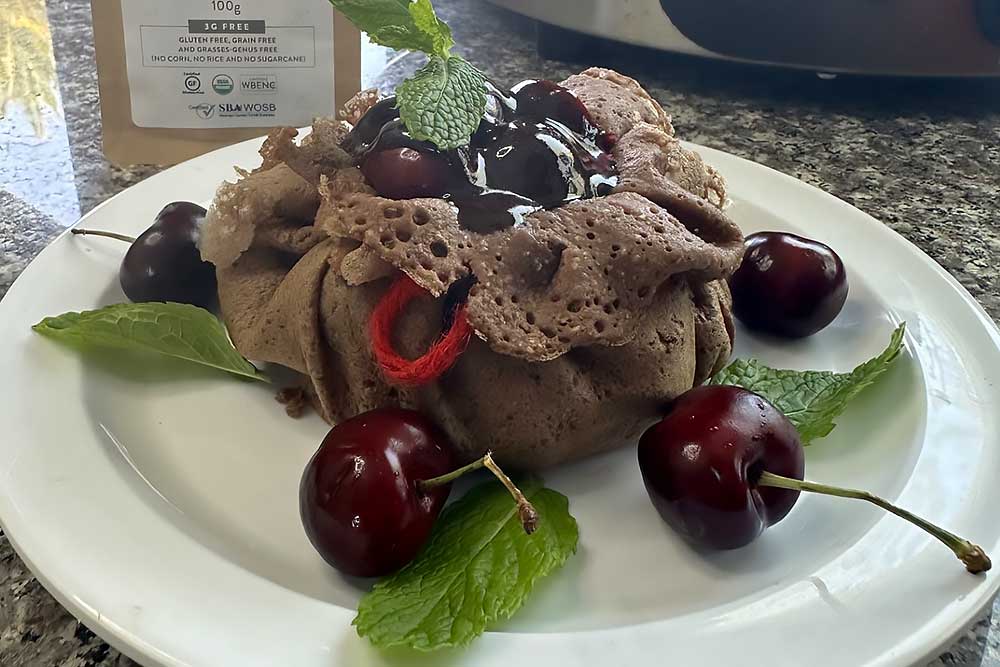 A gluten-free crepe filled with cherries, drizzled with chocolate sauce, and garnished with fresh mint leaves sits on a white plate, surrounded by whole cherries.…| Daré Vita Café | Las Vegas