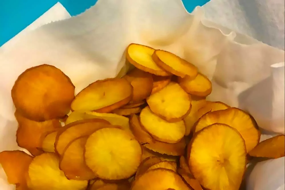 A bowl of golden brown, crispy carrot chips sits on a white paper towel, showcasing a vibrant, healthy snack option. These allergen-friendly chips offer fresh…| Daré Vita Café | Las Vegas