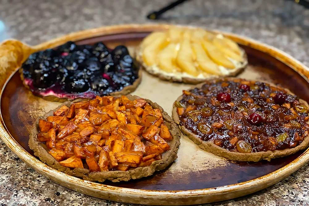 A rustic tray features four vibrant, allergen-friendly tarts: blueberry, pear, apple, and mixed fruit. Each tart showcases fresh, wholesome ingredients, ideal for those with food sensitivities or seeking healthy living.| Daré Vita Café | Las Vegas