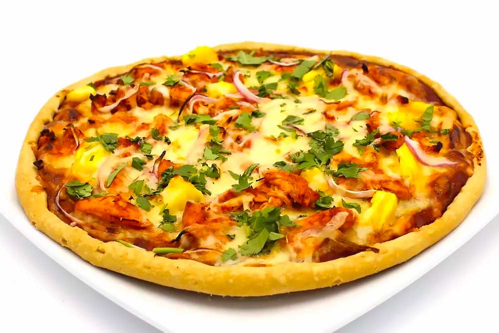 A vibrant, plant-based pizza topped with fresh pineapple, red onions, and cilantro, resting on a white plate. The pizza features a wholesome, allergen-friendly crust, ideal for those seeking healthy and nourishing meals.| Daré Vita Café | Las Vegas