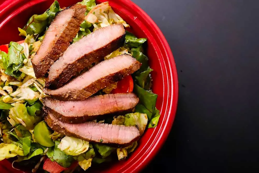 A vibrant salad in a red bowl, topped with slices of grilled meat, surrounded by fresh greens, tomatoes, and cabbage, highlighting fresh flavors and nourishing, wholesome meals made with natural, clean ingredients for healthy living.| Daré Vita Café | Las Vegas