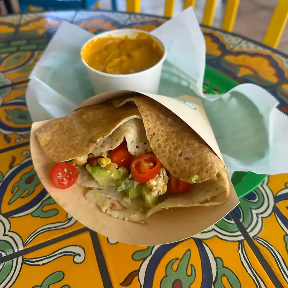 A vibrant, gluten-free crepe filled with fresh produce like cherry tomatoes and avocado, served on a colorful table, showcasing allergen-friendly, nourishing ingredients for healthy living. A cup of orange soup accompanies the dish.| Daré Vita Café | Las Vegas
