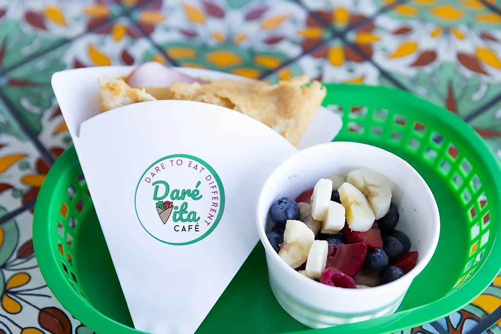 A green tray holds a crepe in a branded paper sleeve with a cup of fresh fruit salad beside it. The dish is allergen-friendly and made from natural, clean ingredients, perfect for those seeking wholesome meals and vibrant, fresh flavors.| Daré Vita Café | Las Vegas