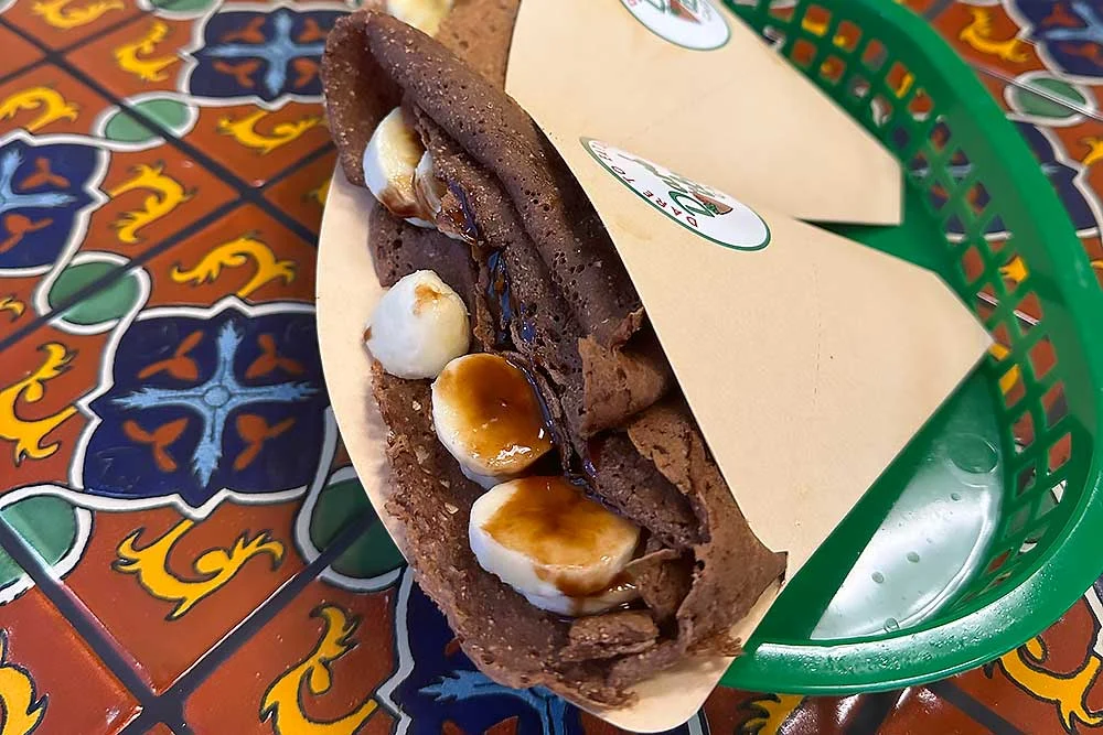 A wholesome, plant-based crepe filled with marshmallows and drizzled with syrup, presented in a green basket on a vibrant, patterned table, showcasing fresh, allergen-friendly ingredients perfect for those seeking healthy living options.| Daré Vita Café | Las Vegas