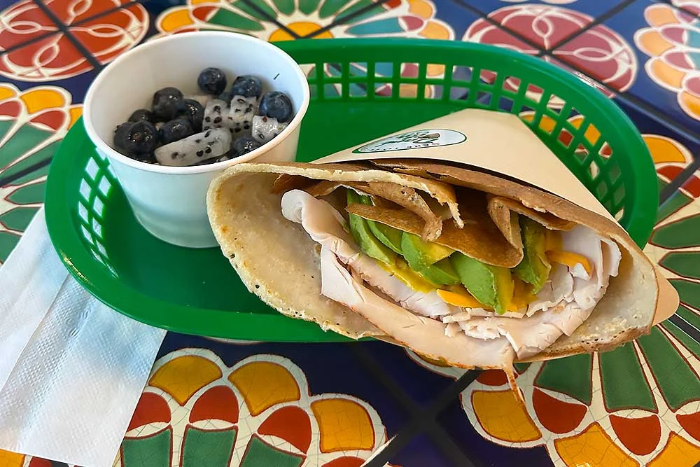A vibrant, allergen-friendly crepe filled with turkey, avocado, and cheese is served in a green basket alongside a cup of blueberries and dragon fruit cubes on a colorful table, highlighting fresh, wholesome ingredients.| Daré Vita Café | Las Vegas