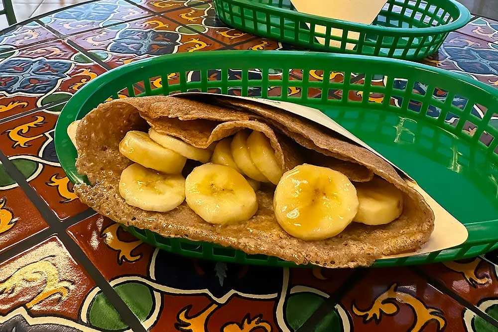 A vibrant, allergen-friendly crepe filled with fresh banana slices is served on a colorful, patterned table. The crepe sits in a green basket, showcasing a healthy, gluten-free and plant-based option, perfect for those with food sensitivities.| Daré Vita Café | Las Vegas