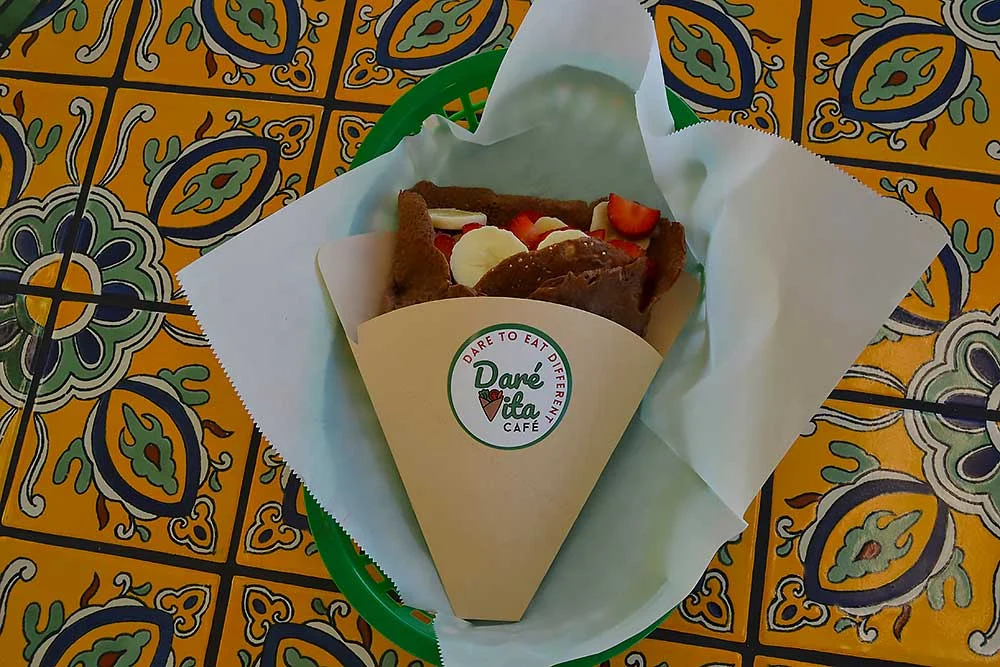 A vibrant crepe from Daré Vita Café, filled with fresh strawberries and banana slices, wrapped in a cone on a decorative tile table. The café focuses on allergen-friendly, gluten-free, and plant-based meals using clean ingredients.| Daré Vita Café | Las Vegas