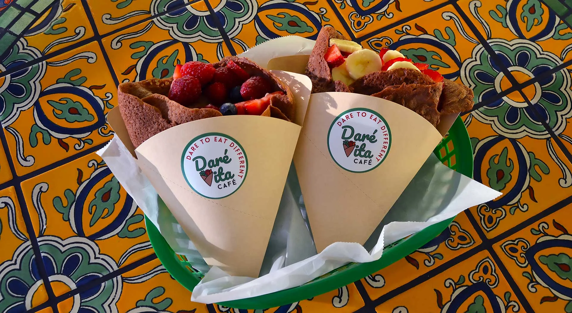 Two brown crepes filled with fresh berries and banana slices are wrapped in paper labeled "Daré Vita Café," set on a vibrant, patterned table. These allergen-friendly treats emphasize fresh, natural ingredients for healthy living.| Daré Vita Café | Las Vegas