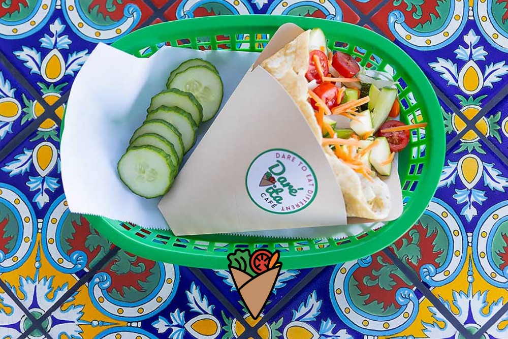 A New Kind of Comfort Food-1 A vibrant green basket holds an allergen-friendly wrap filled with fresh, colorful veggies alongside sliced cucumbers on a decorative blue and yellow tiled table, representing healthy, natural, and fresh produce from Daré Vita Café.| Daré Vita Café | Las Vegas