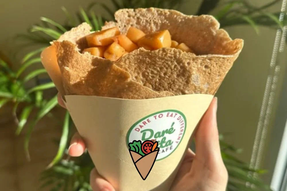 From Crêpe Fridays to Community_ How Food Brings Us Together (4) A hand holds a gluten-free, plant-based crepe filled with fresh fruit at Daré Vita Café. The crepe is wrapped in a branded paper sleeve, emphasizing allergen-friendly and natural ingredients, with a backdrop of green plants.| Daré Vita Café | Las Vegas