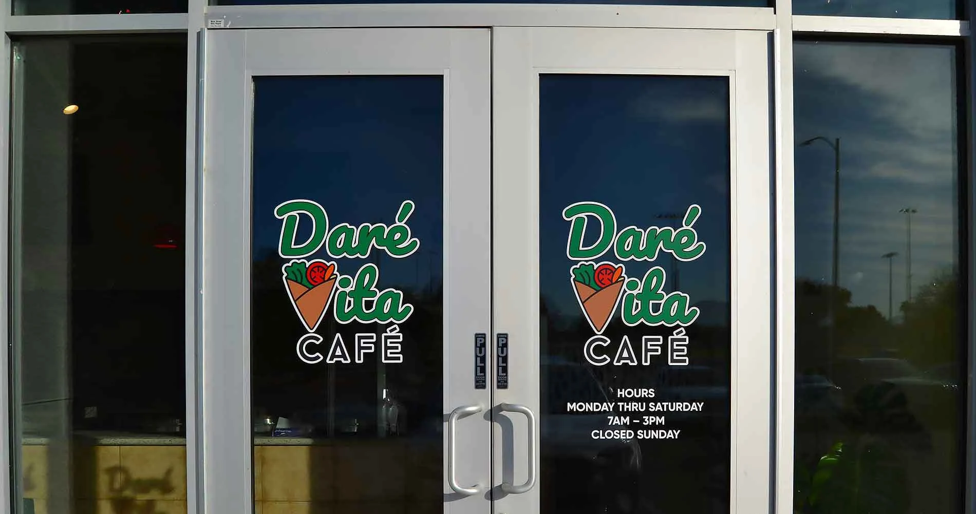 The entrance of Daré Vita Café, featuring vibrant logos with vegetables, promoting allergen-friendly, fresh, and wholesome meals. The glass doors display operating hours and reflect…| Daré Vita Café | Las Vegas