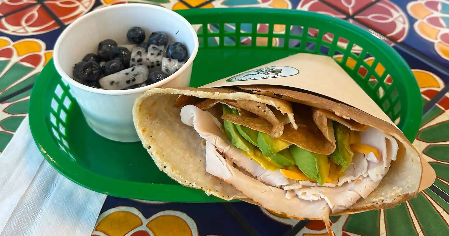 A vibrant wrap with turkey, avocado, and cheese sits in a green basket beside a cup of blueberries and dragon fruit, showcasing allergen-friendly and fresh flavors on a colorful table.| Daré Vita Café | Las Vegas