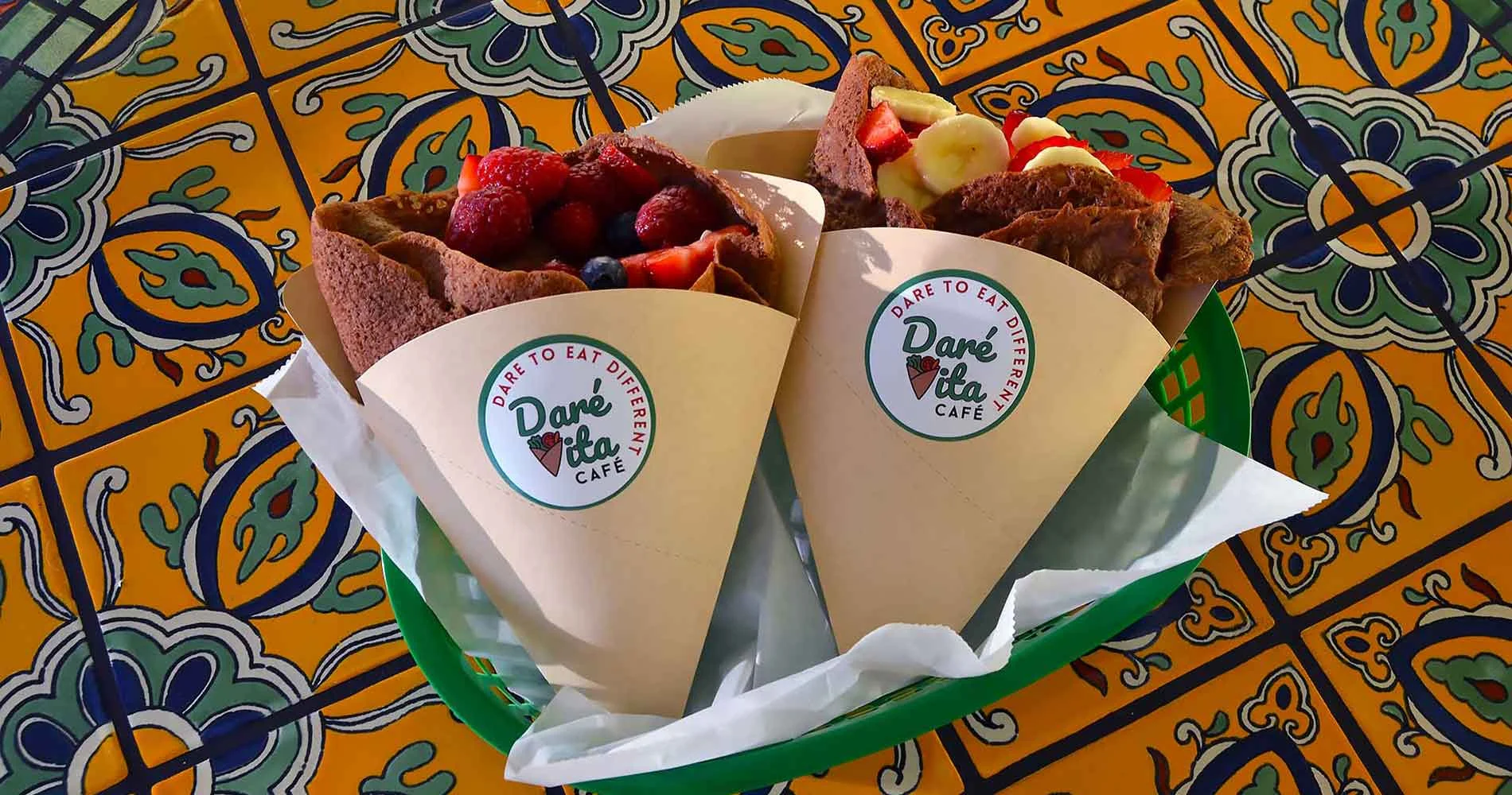 Two gluten-free crepes filled with vibrant fresh fruit, wrapped in paper cones with Daré Vita Café logos, rest in a green basket on a colorful, patterned table.| Daré Vita Café | Las Vegas